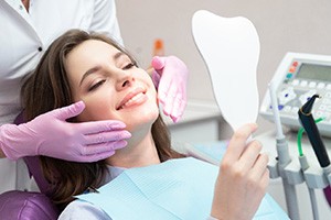 Woman in dental chair smiling into mirror with dentist’s purple-gloved hands near her cheeks