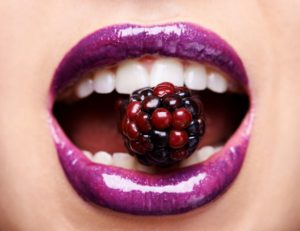 Close up of a mouth with purple lipstick holding a raspberry between the teeth